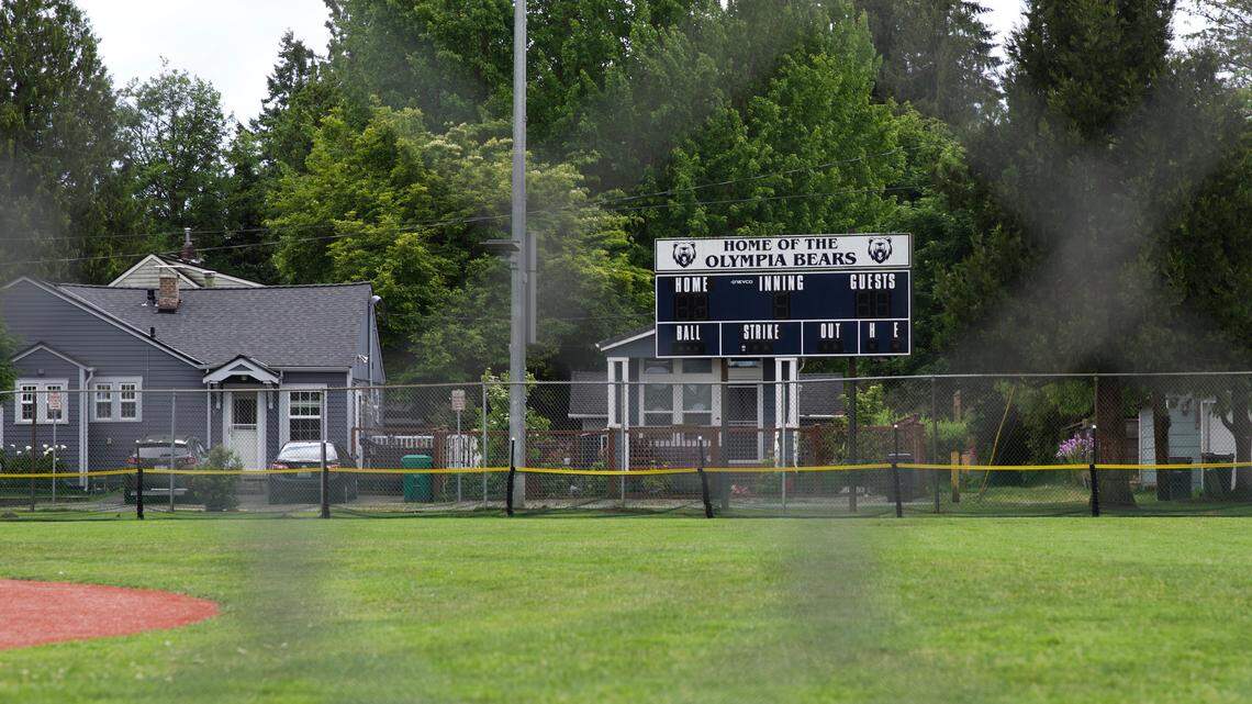 A dark blue scoreboard reading "Home of the Olympia Bears" stands behind a chain link fence in a green field. A gray house stands behind the field.