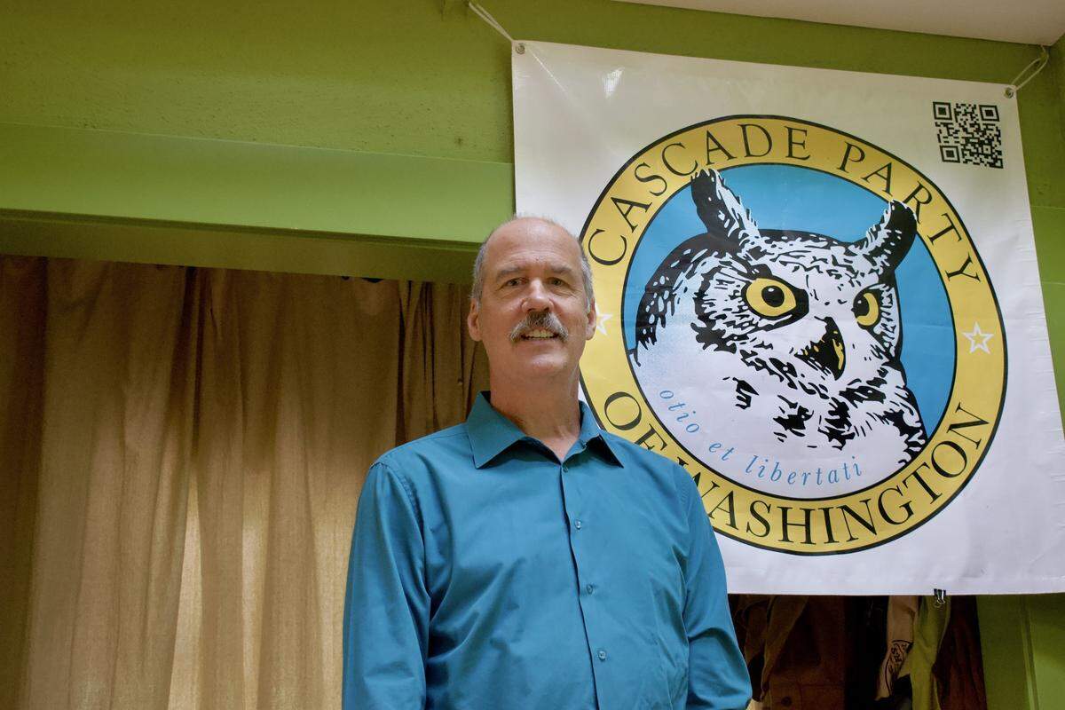 Krist Novoselic stands in front of a Cascade Party banner in his home. The banner features a seal with an owl surrounded by text which reads “Cascade Party of Washington, otio et libertati”.