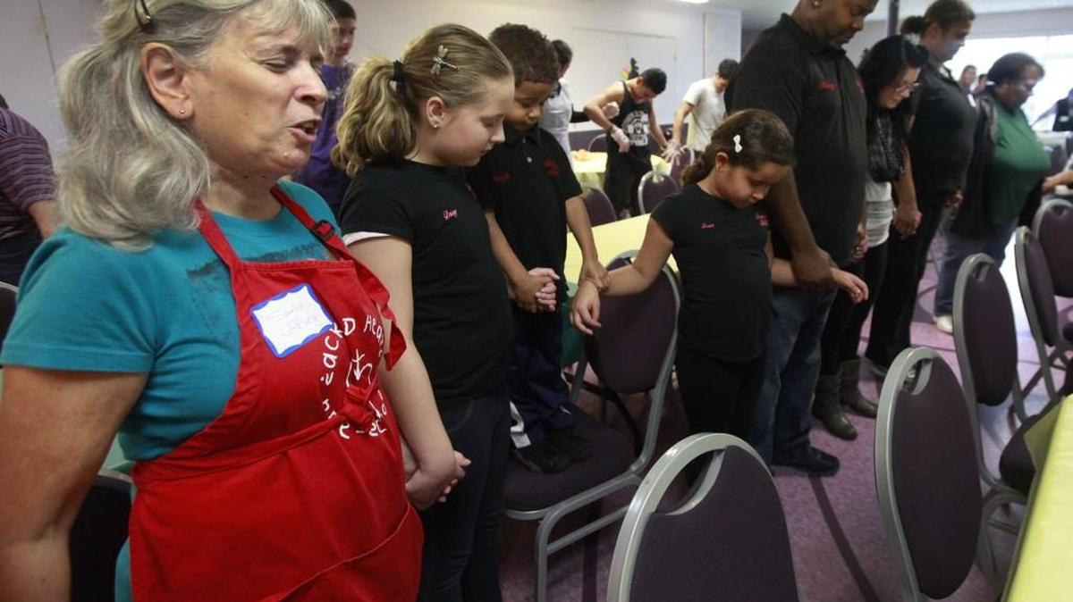 Standing with meal organizer Rodney O’Neill and his children Daisy, Rodney and Grace, volunteer Suellen Apley offers the opening prayer for last year’s Barb O’Neill Family and Friends dinner at The United Churches, downtown Olympia.