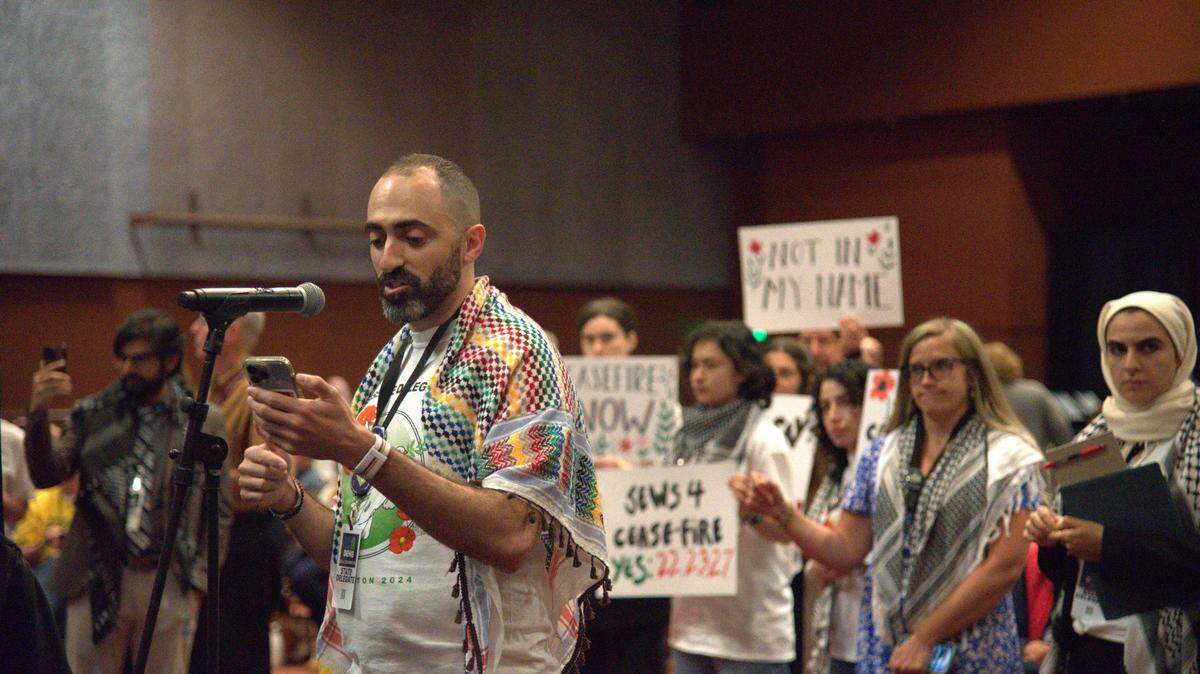A delegate at the Washington State Democratic Convention speaks to resolutions about Israel’s war on Gaza while other delegates hold signs in the background.
